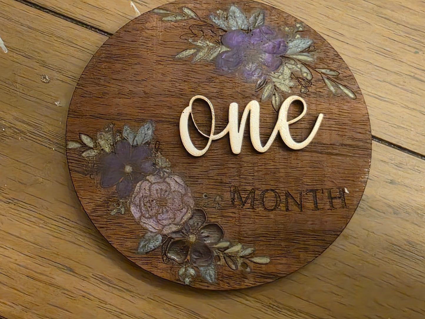Wooden circular milestone with floral designs and 'ONE MONTH' text on a wooden surface